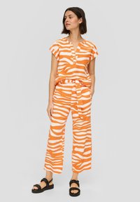 Oranje en wit gestreept twee-delig outfit met een shirt met korte mouwen en brede broekspijpen, met een strik in de taille en platte sandalen.