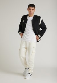 Giacca varsity nera con accenti bianchi sopra una t-shirt bianca, abbinata a pantaloni cargo crema e sneakers bianche, con tasche cargo.