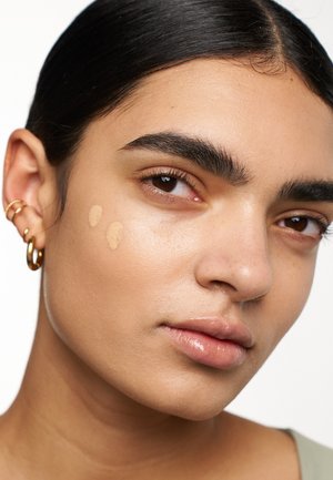 Fond de teint kleurstalen in een beige tint tegen een gladde huid. Model met donker haar, oorringen en een subtiele, natuurlijke afwerking.