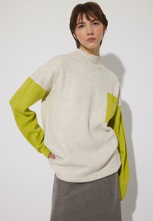 Jeune personne en pull crème avec manches et poche vert citron, portant une jupe grise, debout devant un fond clair uni.