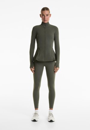 Olijfgroene sportjack met hoge kraag en rits, gecombineerd met bijpassende leggings; beide gemaakt van glad, rekbaar materiaal.