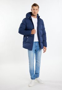 Marineblå polstret jakke med hætte, lynlåslukning, elastiske manchetter og to lynlåslommer i siden, parret med lyseblå jeans og hvide sneakers.