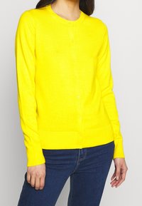 Gelbe Strickjacke aus weichem Strickstoff, mit einem runden Ausschnitt, vorderen Knöpfen und gerippten Bündchen und Saum. Über dunklen Jeans getragen.