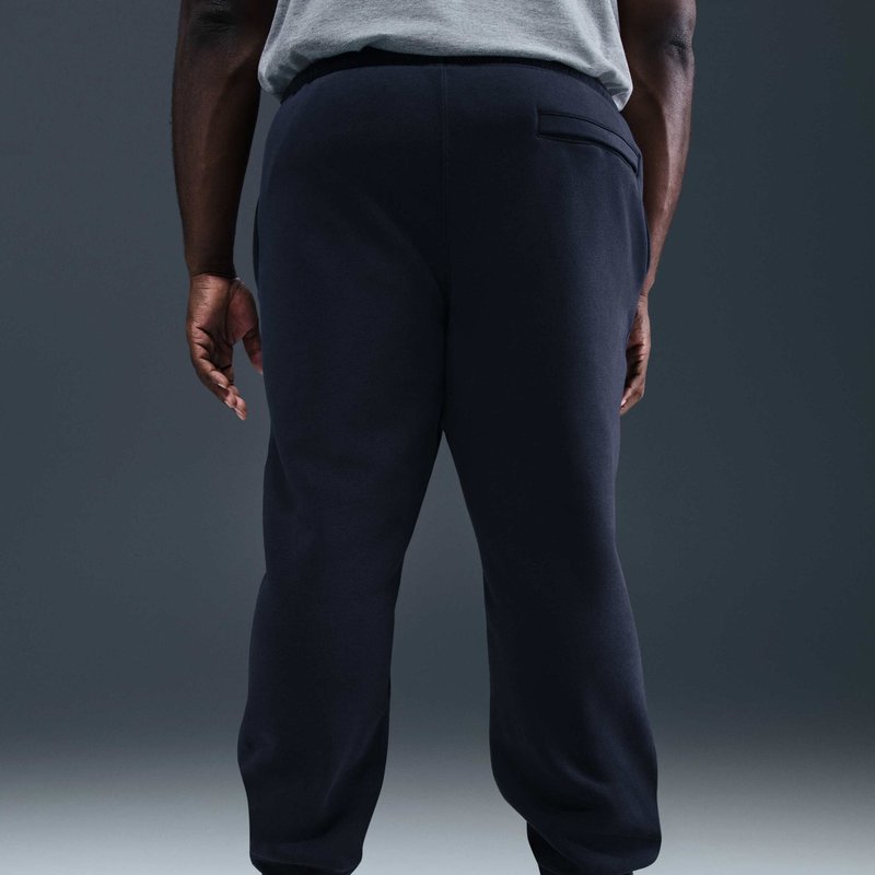 Persona in piedi di spalle alla fotocamera, indossa una maglietta grigia e pantaloni sportivi scuri, mani rilassate ai lati, su uno sfondo neutro.