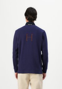 Pólo de manga longa azul-marinho com colarinho preto em contraste, apresentando uma grande letra "H" castanha nas costas e remendos nos cotovelos.