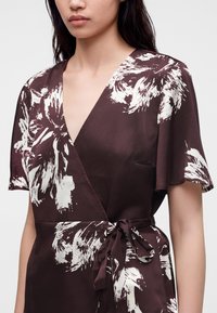 Femme portant une robe portefeuille marron avec des motifs floraux abstraits blancs et des manches courtes flottantes attachées à la taille avec une ceinture en tissu.