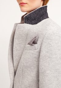 Jasnoszary wełniany płaszcz z ciemnoszarym filcowym wysokim kołnierzem. Posiada małą, teksturowaną poszetkę. Gładka, miękka w dotyku faktura.