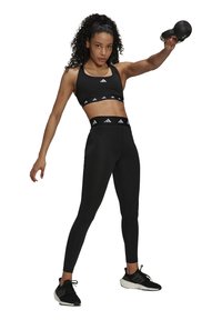 Svart sport-bh och höga tights med vita Adidas-loggor. Modellen håller en kettlebell och bär svarta träningsskor. Lockigt hår synligt.