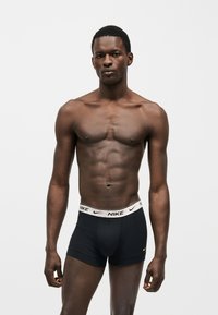 Homme musclé debout face à l'avant, portant un boxer Nike noir avec une ceinture blanche et un petit logo Nike jaune sur la cuisse gauche.