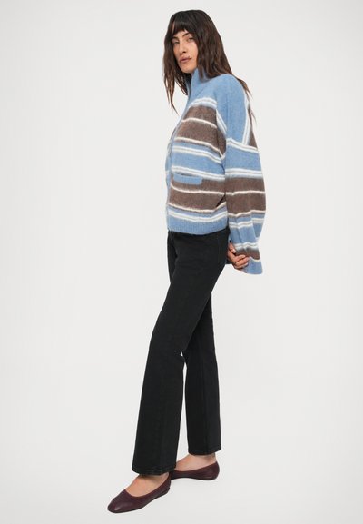 Kvinde stående i sorte jeans, kastaniefarvede flade sko og en blå, brun og hvid stribet fuzzy lynlåstrøje med hænderne bag ryggen.