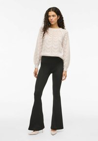Suéter de punto rosa claro con un patrón en forma de chevrón, combinado con pantalones acampanados negros. La modelo lleva zapatos de tacón nude.