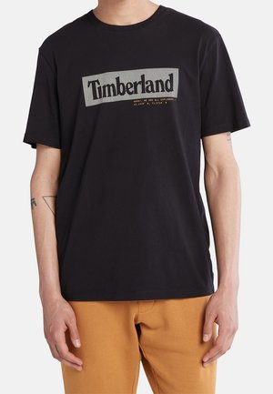 Person trägt ein schwarzes Timberland T-Shirt mit weißem Logotext und hellbrauner Hose, steht mit entspannten Armen an den Seiten.