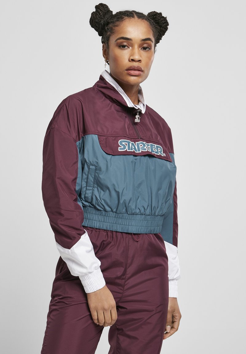 Cropped jacket with a burgundy and teal design. Features elastic waistband, zip closure, and white accents on sleeves. Logo displayed prominently.
