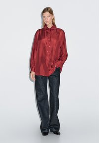 Red satin long-sleeve shirt with a stand collar, featuring a button front. Paired with loose dark denim trousers and black shoes.