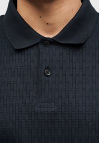 Gros plan sur une personne portant un polo texturé bleu marine foncé avec deux boutons noirs attachés au col.