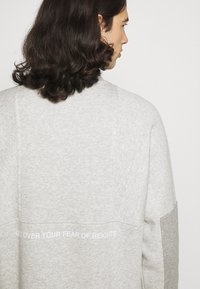 Grå sweatshirt med en blandning av material, med ljus- och mörkgrå paneler; texten "GET OVER YOUR FEAR OF HEIGHTS" i vitt på baksidan.