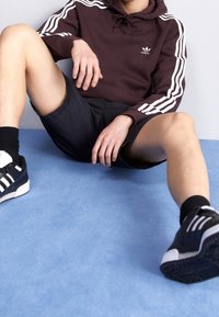 Sudadera con capucha color marrón con rayas blancas en las mangas, pantalones cortos negros y zapatillas deportivas negras con detalles blancos, sentado sobre un fondo azul.