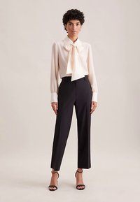 Blusa crema con collo a fiocco, maniche lunghe e pantaloni neri a vita alta. Il modello indossa tacchi neri con cinturini. Tessuto liscio e vestibilità sartoriale.