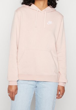 Osoba ubrana w jasnoróżową bluzę Nike z kapturem i kieszenią z przodu oraz jasnoniebieskie dżinsy, z rękami swobodnie opuszczonymi wzdłuż ciała na jednolitym tle.