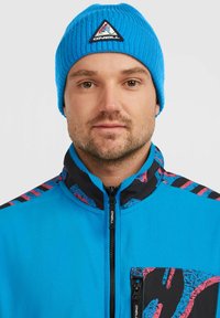 Blå ribbede beanie med et trekantet logo. Jakken har en livlig blå front, sort krave og farverige mønstrede detaljer på skuldrene.