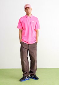 Helles pinkes Polohemd mit kurzen Ärmeln, kombiniert mit lockeren braunen Hosen. Eine hellrosa Kappe und schwarze Sneakers mit blauen Akzenten runden das Outfit ab.