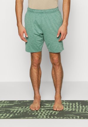 Man staat op blote voeten op een groene yoga-mat met patroon, draagt groene shorts met elastische tailleband en een beige shirt, tegen een effen witte achtergrond.