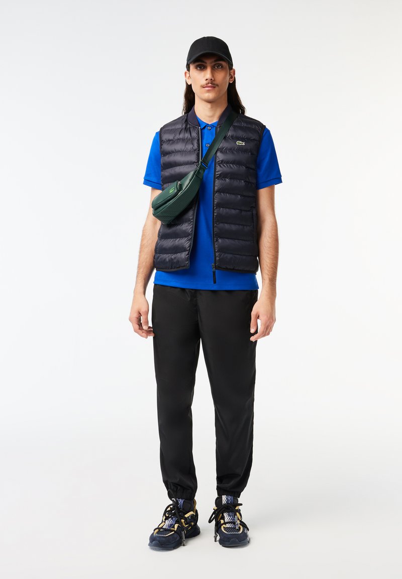 Homme portant une casquette noire, un polo bleu, un gilet matelassé noir, un survêtement noir, des baskets noires et jaunes, et un sac bandoulière vert, se tenant devant un fond blanc.