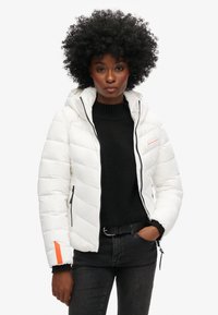 Doudoune blanche avec une capuche, design matelassé, et accent orange sur la manche. Assortie à un pull noir et un jean noir.
