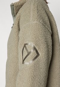 Giacca in pile beige con tessuto testurizzato, chiusura zip davanti e un dettaglio logo argento sulla manica sinistra. Aspetto morbido e accogliente.