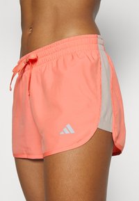 Primer plano de una persona que lleva pantalones cortos deportivos Adidas de color coral brillante con un logo blanco y panel lateral.