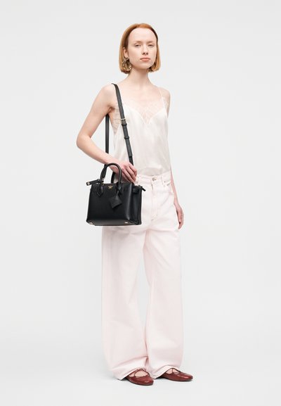 Sac à main en cuir noir avec ferrures dorées, poignées robustes et bandoulière amovible, associé à un débardeur léger transparent et un pantalon large rose pâle.