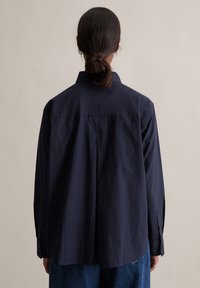 Marineblaues Langarmhemd aus glattem Stoff. Verfügt über eine Rückenpasse und knöpfbare Manschetten, mit lockerem Schnitt und abgerundetem Saum.