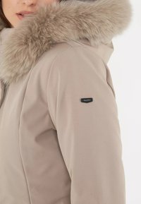 Giacca isolante beige con cappuccio rifinito in pelliccia. Presenta un tessuto morbido, una silhouette aderente e una piccola etichetta con logo sulla manica.