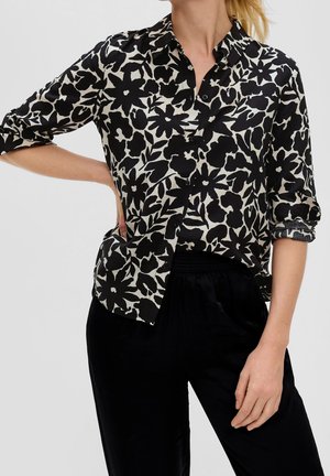 Vrouw draagt een zwart-wit bloemenblouse met opgerolde mouwen en een zwarte broek, poseert met één hand op haar heup.