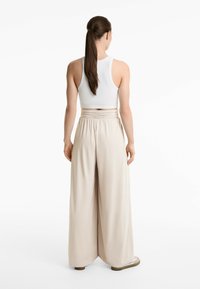 Frau mit langen braunen Haaren in einem Pferdeschwanz, die ein weißes ärmelloses Crop-Top und lockere beige hochgeschnittene weite Hosen trägt, steht mit dem Rücken zur Kamera.