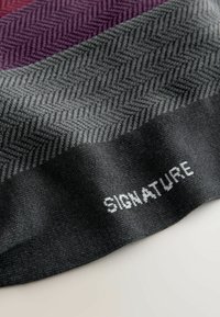 Teksturalny szalik z wzorem w rybie łuski w odcieniach szaro-fioletowych, z widoczną białą metką "SIGNATURE" naszytą na tkaninie.