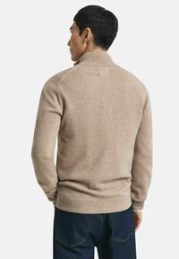 Man die wegkijkt, gekleed in een lichtbruine coltrui met hoge kraag en donkerblauwe jeans, tegen een egale witte achtergrond.