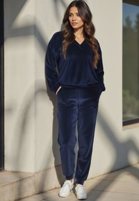 Marineblauer Velour-Trainingsanzug mit einem V-Ausschnitt-Oberteil und schmal zulaufenden Hosen mit gerippten Bündchen, kombiniert mit weißen Sneakern. Das Gesicht ist nicht sichtbar.