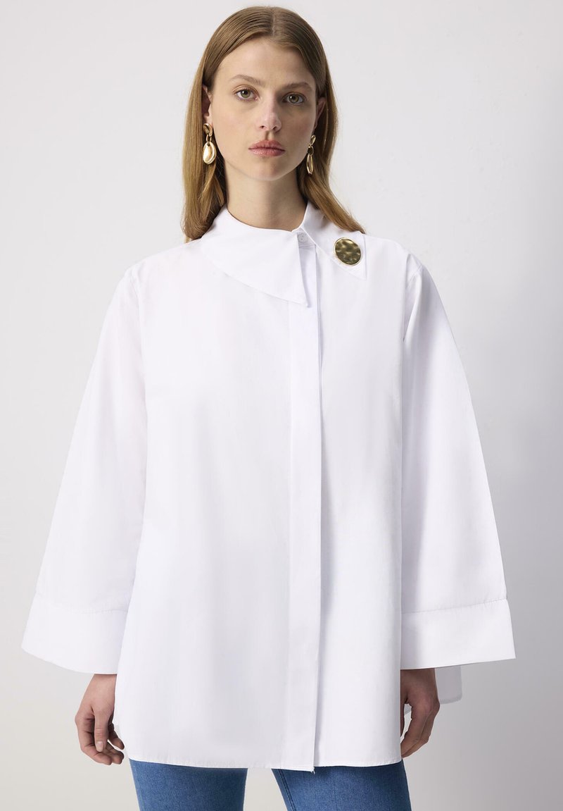 Femme portant une chemise blanche oversize boutonnée avec des manches larges et une grande broche ronde en or sur le col, associée à un jean bleu.