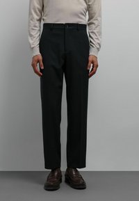 Pantalons noirs sur mesure à coupe droite, avec une texture lisse, des passants pour ceinture et une fermeture à un bouton. Portés avec des mocassins marron.