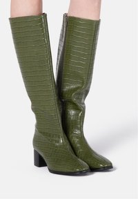 Bottes hauteur genou en simili cuir vert brillant avec texture crocodile, dotées d'un bout carré et d'un talon bloc. Détail zippé sur le côté.