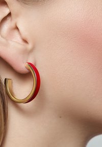 Goldene Creole mit rotem Emaille-Äußeren. Glatte Oberfläche, kreisförmiges Design, mittlere Größe, im Ohr getragen, das den Farbenkontrast zur Geltung bringt.