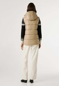Beige gewatteerde vest met een capuchon, geruite textuur en verstelbare trekkoorden. Draag het over een zwarte trui met witte accenten, gecombineerd met witte broeken.