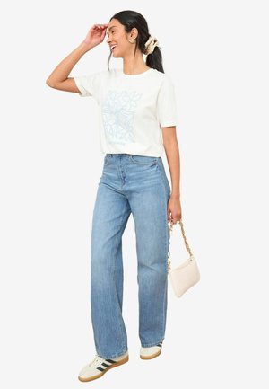 T-shirt blanc avec un graphique floral bleu, associé à un jean large bleu clair et des baskets blanches avec des accents verts. Tenant un petit sac à main crème.