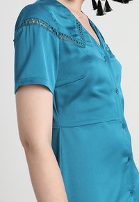 Teal short-sleeve blouse with a button-down front, featuring intricate lace detailing along the shoulders and a smooth, satin-like texture.