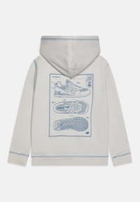 Gaiši pelēks hoody ar zilu shēmu printu, kas attēlo sporta apavi aizmugurē, ar kontrastējošām šuvēm un kapuci ar auklu.