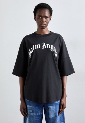 Femme portant un t-shirt noir ample avec le texte « Palm Angels » et un jean bleu avec imprimé de journaux, se tenant devant un fond clair.