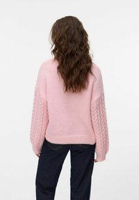 Pull en tricot rose avec des manches en torsades, coupe décontractée, ourlet côtelé et texture douce. Porté avec un pantalon en denim foncé.