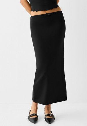 Femme portant une jupe noire ajustée longueur cheville avec un petit détail de nœud à la taille et des ballerines noires à bout pointu avec des lanières cloutées.