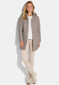 Lichtgrijze pufferjack met verticale stiksels, ritssluiting en capuchon, gelaagd over een witte top, gecombineerd met beige broeken en schoenen.